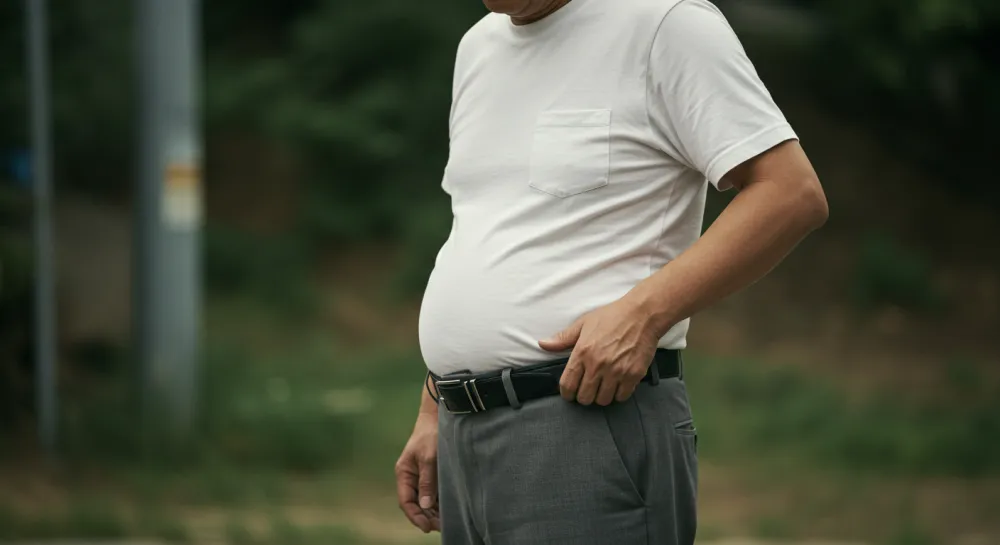 배가 나온 중년 남성이 허리띠를 잡고 있는 모습. 복부 비만의 위험성을 시각적으로 보여줌.